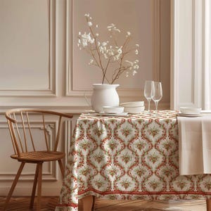 Könnte beinhalten: Ein gedeckter Esstisch. Der Tisch ist mit einer gemusterten Tischdecke bedeckt, die ein sich wiederholendes Design aus roten Beeren und grünen Blättern auf cremefarbenem Hintergrund aufweist. Eine weiße Vase enthält weiße Blumen. Weingläser und weiße Gerichte sind auf dem Tisch.
