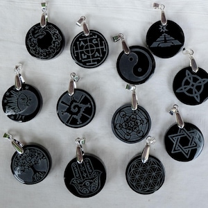 Op de afbeelding: Een verzameling zwarte, ronde hangers met zilverkleurige beugels. Elke hanger heeft een ander geëtst ontwerp, waaronder een boom, een hand, een ster, een yin-yang symbool en andere geometrische en symbolische patronen. De hangers worden gepresenteerd op een wit oppervlak.
