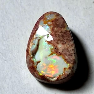 Pode incluir: Uma pedra preciosa de opala boulder em forma de lágrima, apresentando uma matriz castanha terrosa texturizada com manchas incrustadas de opala iridescente. A opala exibe flashes vibrantes de luz verde, laranja e amarela sobre um fundo branco leitoso.