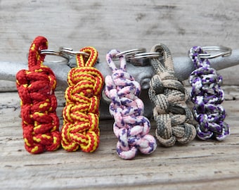 May include: Five handmade keychains in various colors. The keychains are made of braided rope and feature a silver key ring. Colors include red, yellow, pink, purple, and camouflage.