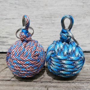 May include: Two decorative ball keychains. One is woven with red, white, and blue cord, and the other with shades of blue and white. Each keychain has a silver-colored ring and a blue loop.