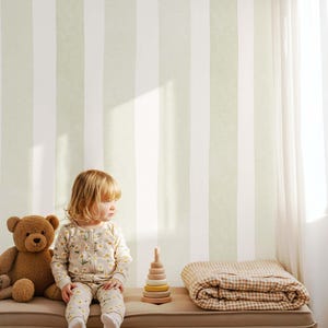 May include: A young child in a floral-print onesie sits on a bench next to a teddy bear, a wooden stacking toy, and a folded blanket. The background features vertical stripes of white and light green. A woven basket is visible.