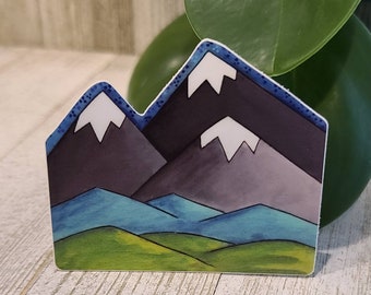 May include: A sticker featuring a stylized mountain range design. The mountains are depicted in shades of gray and purple, with white snow caps. The foreground shows green and blue rolling hills. The sticker has a blue border with white dots.