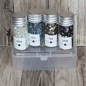May include: Four small glass jars with silver lids filled with different colored rhinestones. The jars are labeled with numbers: 3024, 413, 762, and 3799. The jars are sitting in a clear plastic storage box.