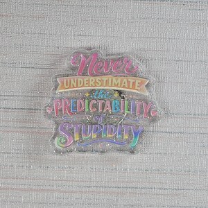 May include: A clear, glittery pin with the text "Never Underestimate the Predictability of Stupidity" in pink, blue, and yellow lettering. The pin has a whimsical, playful design.