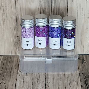 May include: Four small glass jars filled with different shades of purple and blue glitter. The jars have silver lids and labels with numbers: 211, 208, 3746, and 550.
