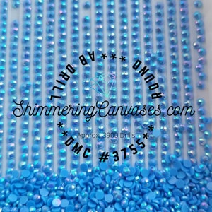 Puede incluir: Primer plano de piedras de imitación de diamante redondas de color azul claro. Las piedras de imitación de diamante están dispuestas en filas y esparcidas sobre una superficie blanca. El texto "Shimmering Canvases.com *DMC #3755* Approx. 3900 Drills ROUND *** AB DRILL" está impreso en la superficie.