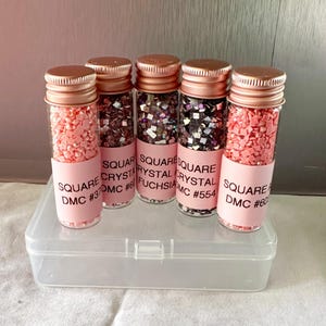 May include: Five small glass jars with rose gold lids filled with pink and iridescent square crystals. The jars are labeled with the text "SQUARE CRYSTAL DMC #31", "SQUARE CRYSTAL DMC #61", "SQUARE CRYSTAL FUCHSIA", "SQUARE CRYSTAL DMC #554", and "SQUARE CRYSTAL DMC #60".