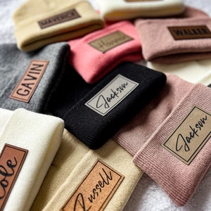 May include: A collection of knitted beanies with personalized leather patches. The beanies are in various colors, including beige, pink, gray, black, and white. The patches feature names such as Gavin, Jackson, Cole, Russell, Walker, and Harper.