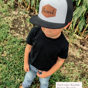 May include: A white and black trucker hat with a brown leather patch that says "Maverick" in cursive. The patch is a hexagon shape and is stitched on.