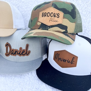 May include: Three different baseball caps with personalized leather patches. The first cap is camouflage with a brown patch that says "Brooks Allen". The second cap is gray with a black patch that says "Daniel". The third cap is black and white with a brown patch that says "Maverick".
