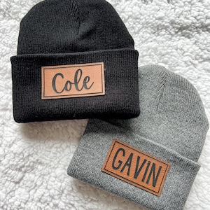 May include: Two knit beanies with personalized leather patches. The beanie on the left is black with a brown leather patch that says "Cole". The beanie on the right is gray with a brown leather patch that says "Gavin".