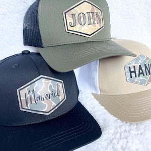 May include: Three trucker hats with personalized name patches. One hat is olive green with a black mesh back and the name "JOHN". Another is black with "Maverick". The third is tan with a white mesh back and "HAN".