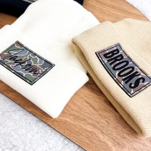 May include: Three knit beanies with personalized patches. The beanies are black, white, and beige. The patches have camouflage designs and the names Cole, Jackson, and Brooks.