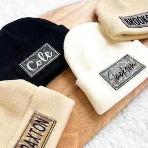 May include: Four beige knit beanies with personalized patches. The patches feature camouflage print and the names "Cole", "Jackson", "Braxton", and "Brooks".