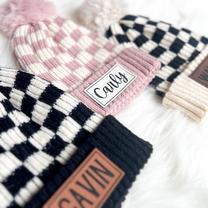 May include: Two black and white checkered knit beanies with pom poms and personalized leather patches. One beanie has a pink and white striped knit beanie underneath. The patches read "Gavin" and "Carly".
