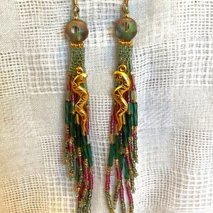 May include: A pair of dangle earrings featuring gold-tone metal hooks, iridescent round beads, and long tassels composed of multi-colored glass seed beads in shades of green, pink, and gold, accented with a gold-tone metal seahorse charm.