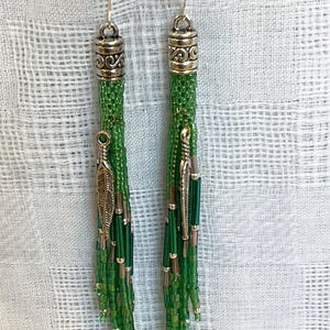 May include: Green beaded tassel earrings with silver-tone accents. Each earring features a decorative silver cap and a dangling feather charm. The earrings are set against a white, textured background.
