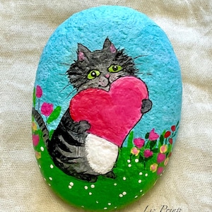 May include: Hand-painted rock art featuring a gray cat holding a pink heart. The cat has green eyes and is set against a blue sky and green grass with pink and yellow flowers. The artist's signature is visible.