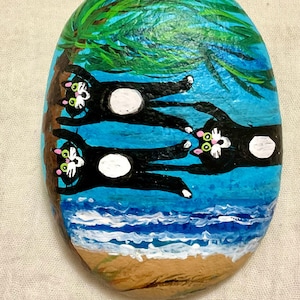 May include: Hand-painted rock art featuring three black cats with white bellies, lounging on a beach under a palm tree. The artwork is set against a blue sky and ocean backdrop, with the artist's signature at the bottom.