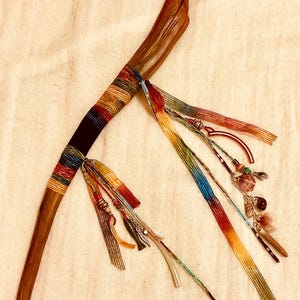 May include: A polished wooden wand featuring a handle wrapped in multi-colored thread. Attached to the handle are several long, gradient-dyed ribbons in shades of red, orange, yellow, and blue, accented with small beads, charms, and feathers.