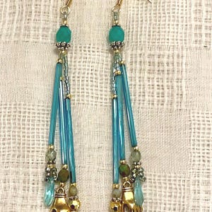 May include: A pair of dangle earrings with gold-colored hooks. Each earring features a turquoise bead, blue glass rods, small beads, gold-colored hand charms, and teardrop-shaped turquoise beads. The earrings are set against a textured white background.