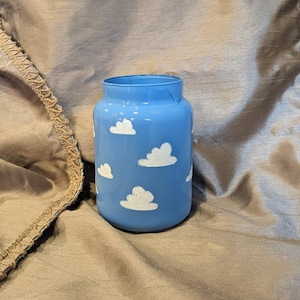 May include: A blue glass jar with white cloud designs. The jar has a wide opening and a cylindrical shape. The clouds are painted in a whimsical style, creating a sky-like appearance. This decorative jar can be used for storage or display.