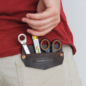 May include: A brown leather pocket organizer with the text "WORK HARD" embossed on it, holding a wrench, a level, and scissors. The organizer is attached to the back pocket of khaki pants. The person is wearing a red shirt.