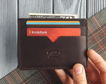 May include: A dark brown leather cardholder with a minimalist design. The wallet holds a few credit cards and a paper bill. The KredoBank logo is visible on one of the cards. The wallet is held against a gray and plaid background.