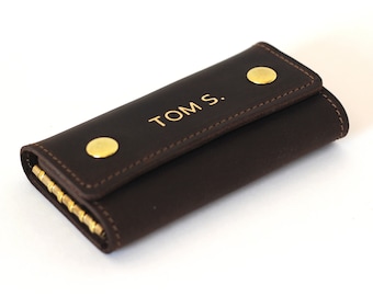Pode incluir: Um porta-chaves de couro castanho com detalhes dourados e o nome "TOM S." gravado na frente.