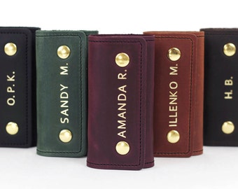 May include: A collection of personalized leather key holders in various colors, including black, green, and burgundy. Each holder is embossed with a name or initials in gold lettering and secured with a gold snap closure.