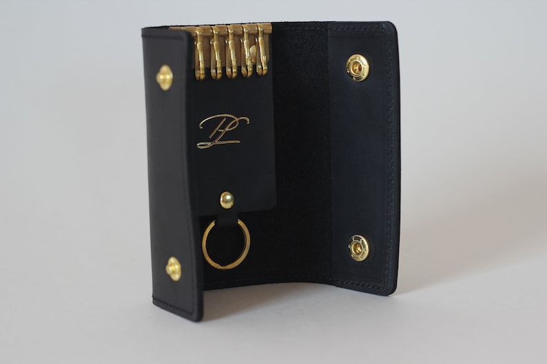 May include: Black leather key holder with a gold ring and a gold monogrammed initial "HL". The key holder has a flap that opens to reveal a key chain with multiple gold key rings.