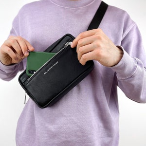 May include: A black leather fanny pack with a zipper closure and a white logo that reads "POL LEATHER STUDIO". A green phone is being placed into the pack.