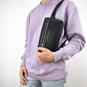 May include: A black leather fanny pack with a silver zipper and the text "Paul Leather Studio" printed on the front. The fanny pack is worn by a person wearing a light purple sweatshirt.