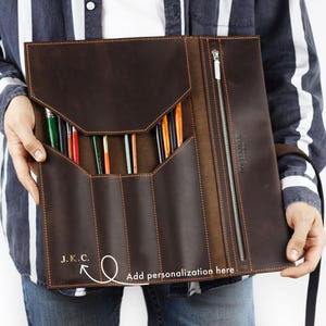May include: A brown leather art supply portfolio with a zipper closure and multiple pockets. The portfolio is filled with various paint brushes and pencils. The text "J.K.C." is visible on the front, with an arrow pointing to the words "Add personalization here".
