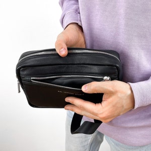May include: A black leather crossbody bag with a zipped pocket and a silver zipper pull. The bag has a black strap and the text "POL LEATHER STUDIO" is printed on the front.