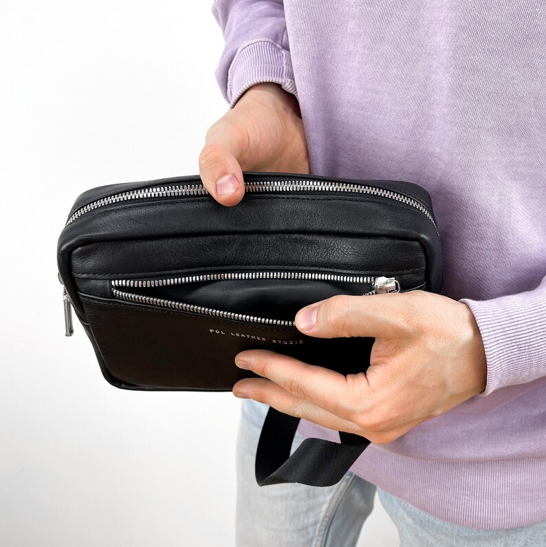 May include: A black leather crossbody bag with a zipped pocket and a silver zipper pull. The bag has a black strap and the text "POL LEATHER STUDIO" is printed on the front.