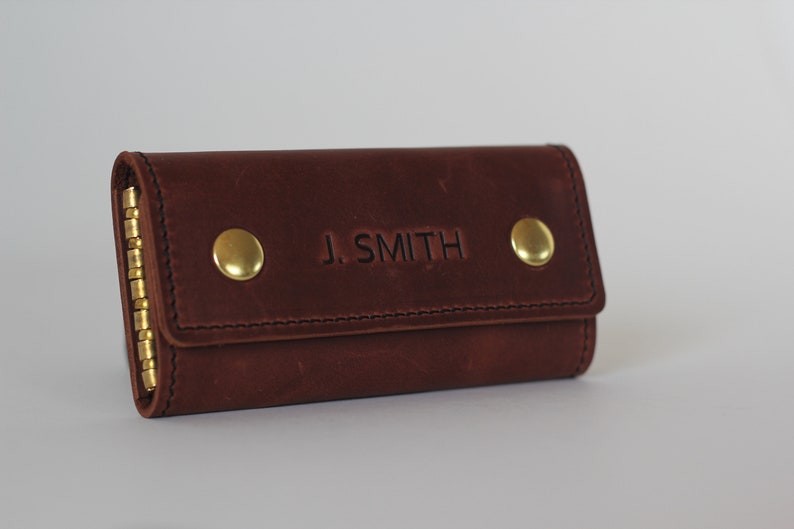 May include: A brown leather key holder with a brass chain and two brass buttons. The key holder is embossed with the name "J. SMITH".