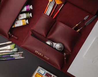 May include: A burgundy leather artist's portfolio case, open to show art supplies. The case contains paint tubes, brushes, palette knives, and a watercolour palette. The text "STARA FOX" is visible on the case.