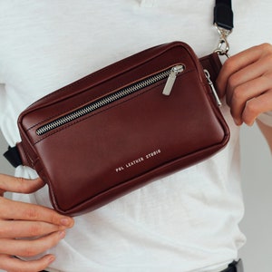 May include: A brown leather fanny pack with a silver zipper and a black strap. The bag has the text "POL LEATHER STUDIO" printed on the front.
