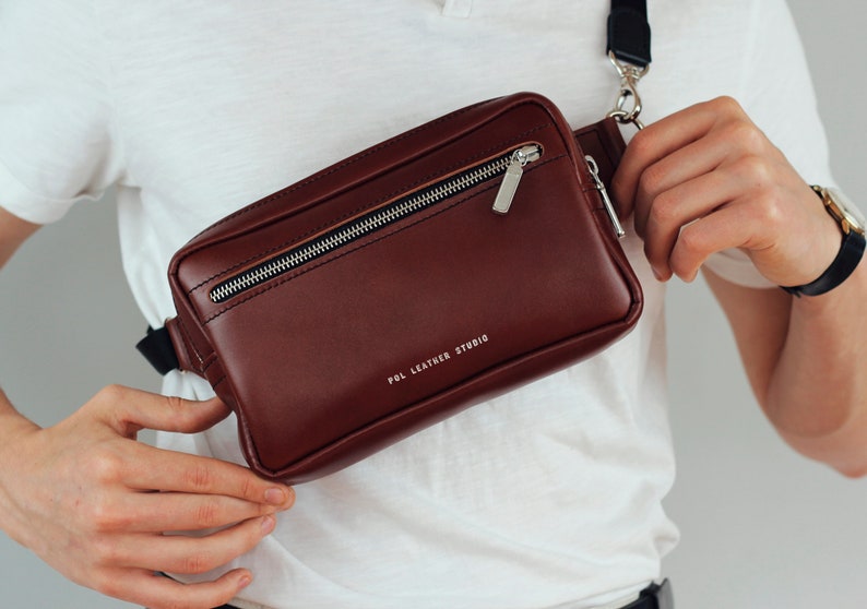May include: A brown leather fanny pack with a silver zipper and a black strap. The bag has the text "POL LEATHER STUDIO" printed on the front.