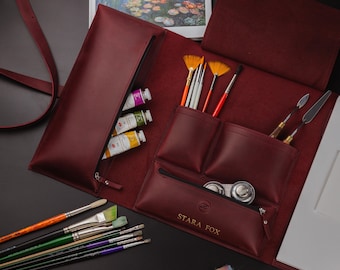 May include: A burgundy leather artist's portfolio, open to display art supplies. It holds paint tubes, brushes, palette knives, and has a zippered pocket. The text "STARA FOX" is visible on the front.