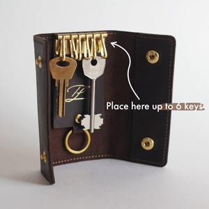 May include: Brown leather key holder with a brass key ring and six key slots. The key holder is open and shows the inside with three keys attached to the ring. The text "Place here up to 6 keys" is visible on the right side of the image.