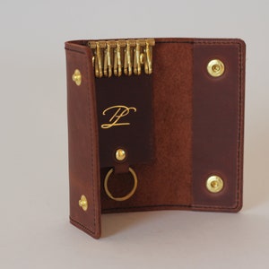 May include: A brown leather key holder with a gold key ring and a gold metal snap closure. The key holder has a small pocket with the letter "L" embossed in gold.