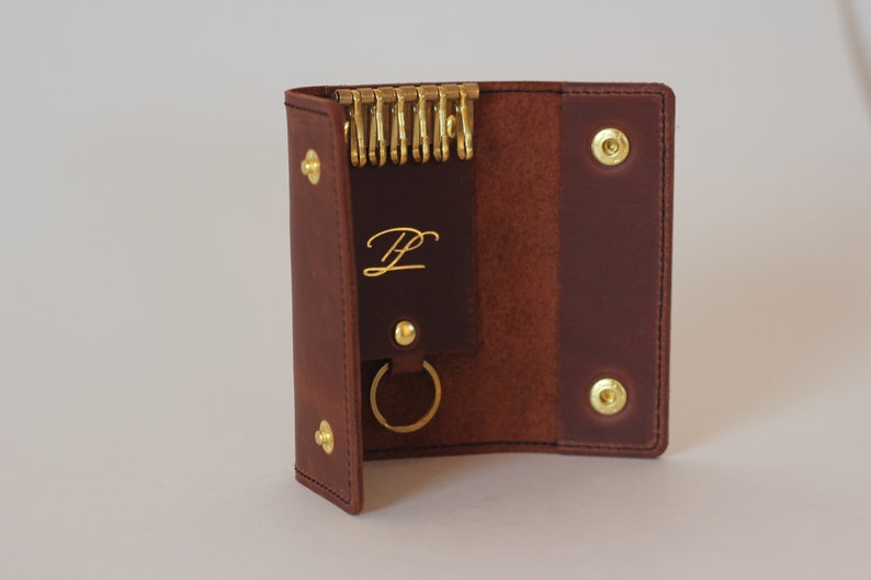 May include: A brown leather key holder with a gold key ring and a gold metal snap closure. The key holder has a small pocket with the letter "L" embossed in gold.