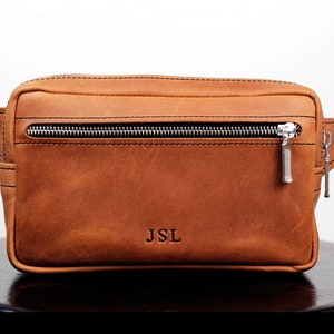 May include: A brown leather fanny pack with a zipper closure and a silver zipper pull. The fanny pack has a black strap and the initials "JSL" are embossed on the front.