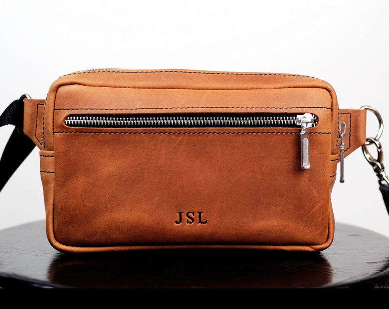 May include: A brown leather fanny pack with a zipper closure and a silver zipper pull. The fanny pack has a black strap and the initials "JSL" are embossed on the front.