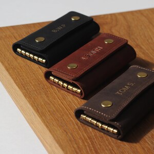 May include: Three leather keychains with personalized names. The keychains are black, brown, and dark brown. Each keychain has a brass snap closure and a brass key ring.