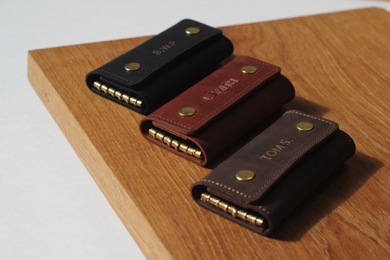 May include: Three leather keychains with personalized names. The keychains are black, brown, and dark brown. Each keychain has a brass snap closure and a brass key ring.
