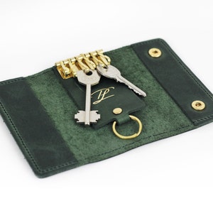 Könnte beinhalten: Ein dunkelgrüner Leder-Schlüsselhalter mit goldfarbener Hardware und zwei Schlüsseln. Der Schlüsselhalter ist offen und zeigt das Innere und eine goldfarbene Initiale. Der Schlüsselhalter wird mit goldfarbenen Druckknöpfen verschlossen.